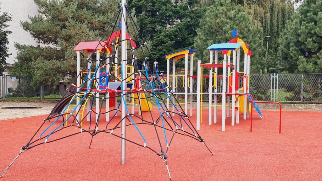 Kolorowy plac zabaw z konstrukcją do wspinania, w tle tor zręcznościowy.